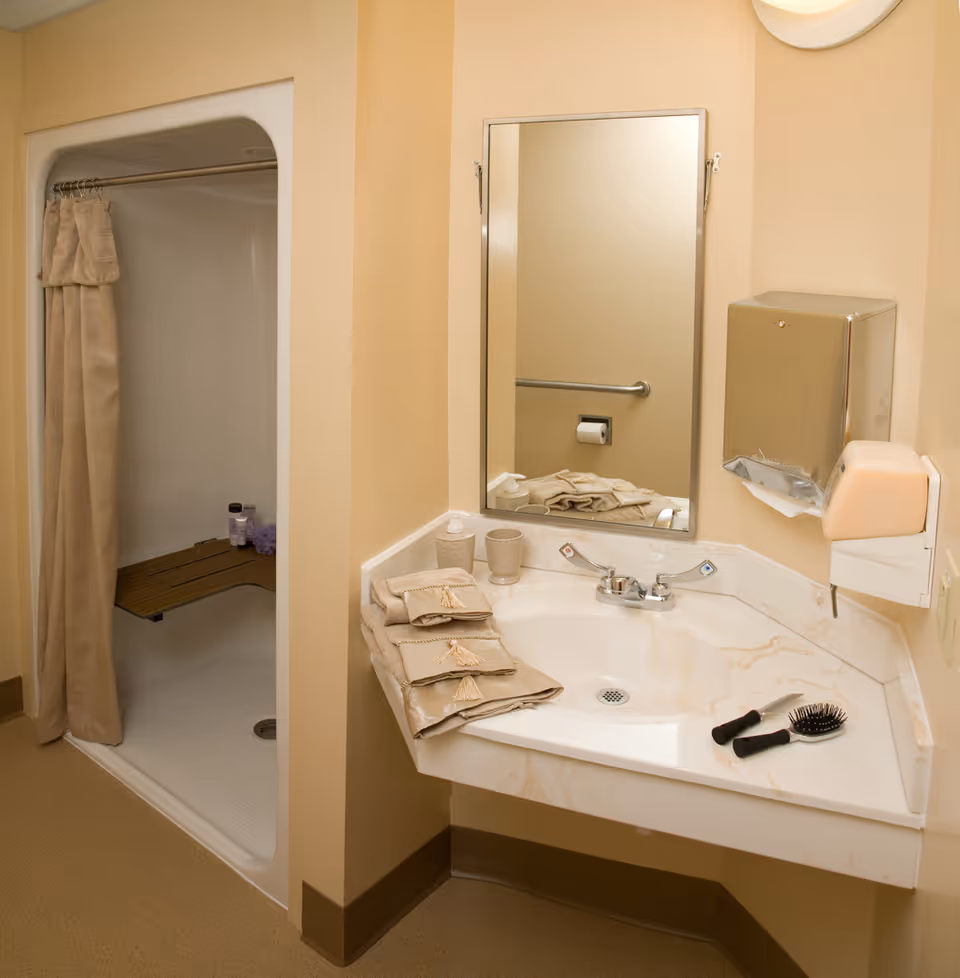 A bathroom with a beige color scheme featuring a corner sink with a marble countertop. On the countertop are folded beige towels, a soap dispenser, a cup, a hairbrush, and a comb. Above the sink is a rectangular mirror. To the left is a walk-in shower with a beige curtain and a wooden bench inside. A metal paper towel dispenser and a soap dispenser are mounted on the wall to the right of the sink.
