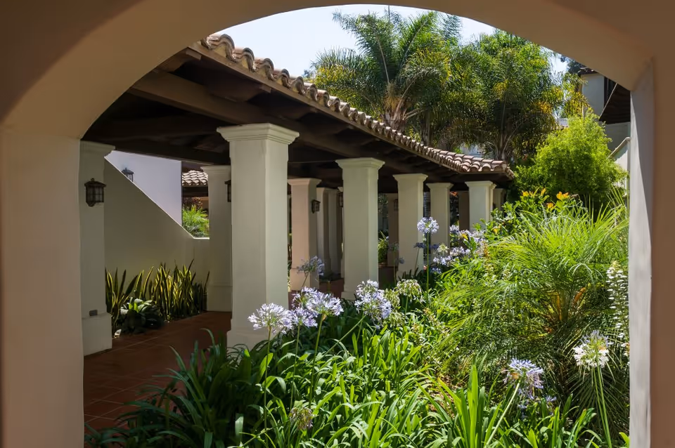 View through an archway of a covered walkway with white columns and a tiled roof, surrounded by lush green plants and flowering bushes in a garden area with palm trees in the background.