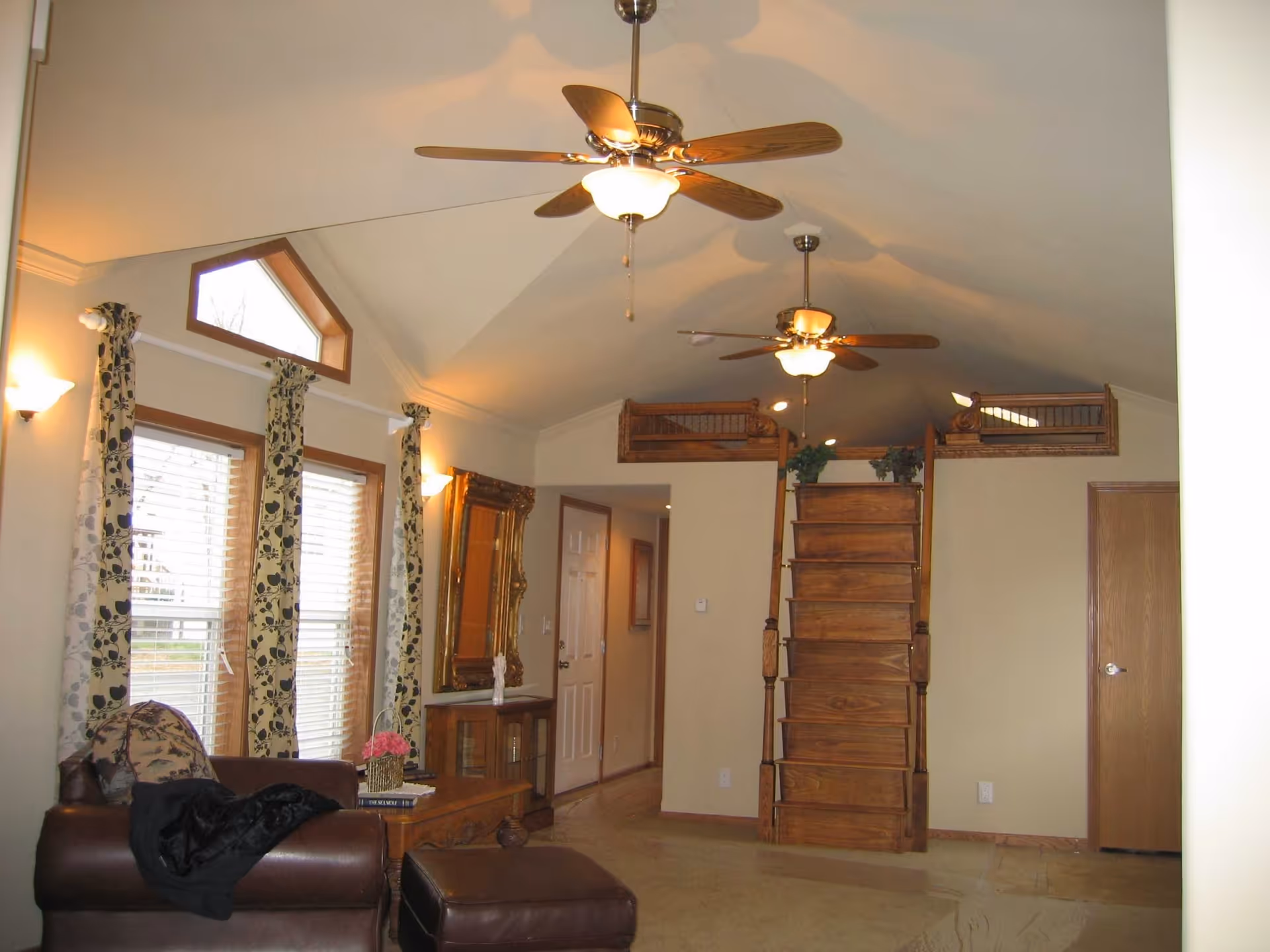 Spacious living room with a vaulted ceiling, two ceiling fans, large windows with patterned curtains, leather seating and a wooden loft reached by a stair.
