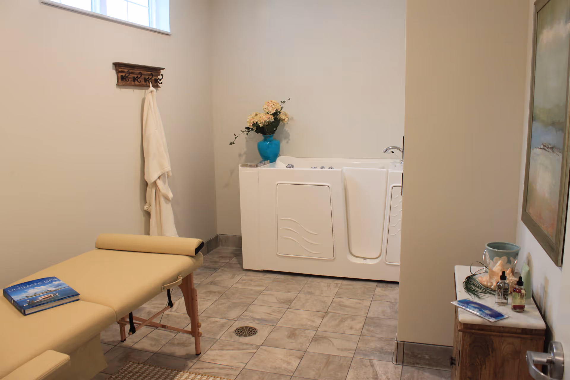 A room with a beige massage table, a white walk-in bathtub, a wooden coat rack with a white robe hanging, a small wooden cabinet with decorative items, and a window letting in natural light.