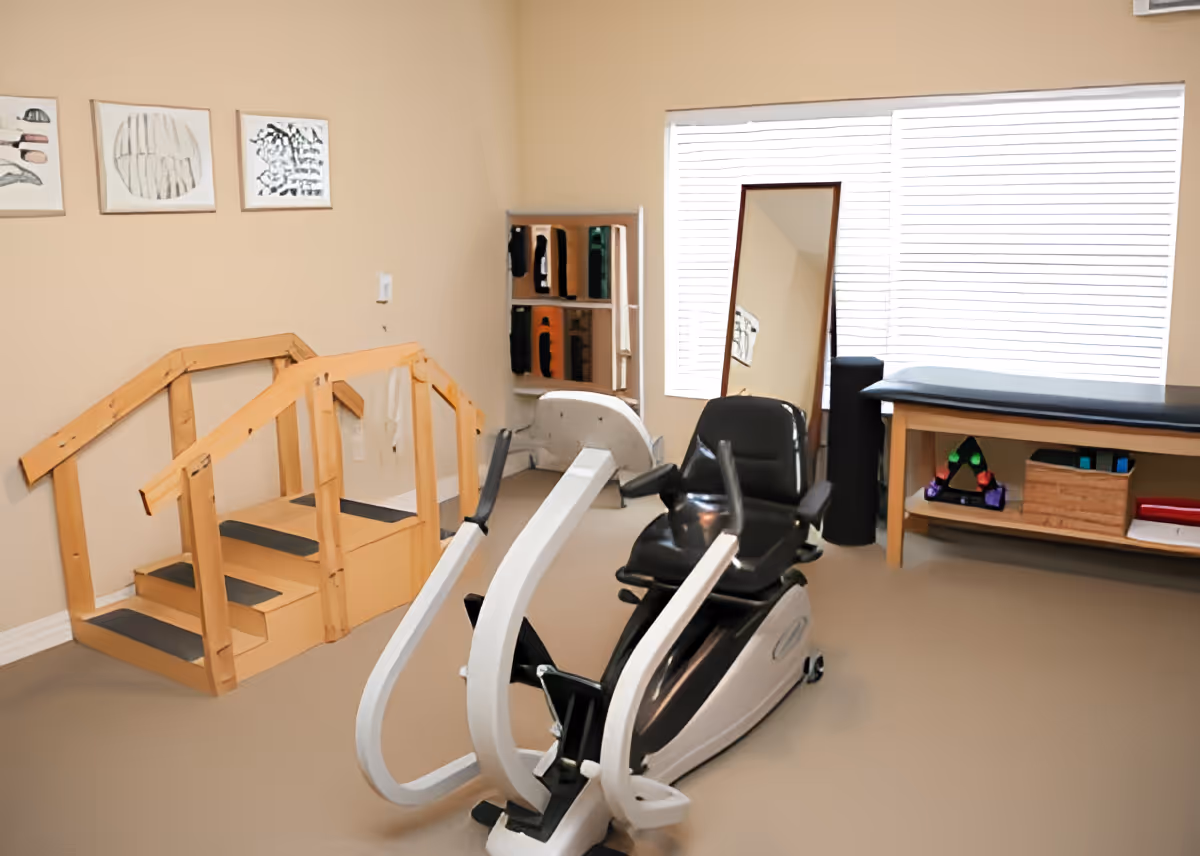 Physical therapy room with a recumbent exercise bike, wooden therapy stairs, a treatment table, and a standing mirror.