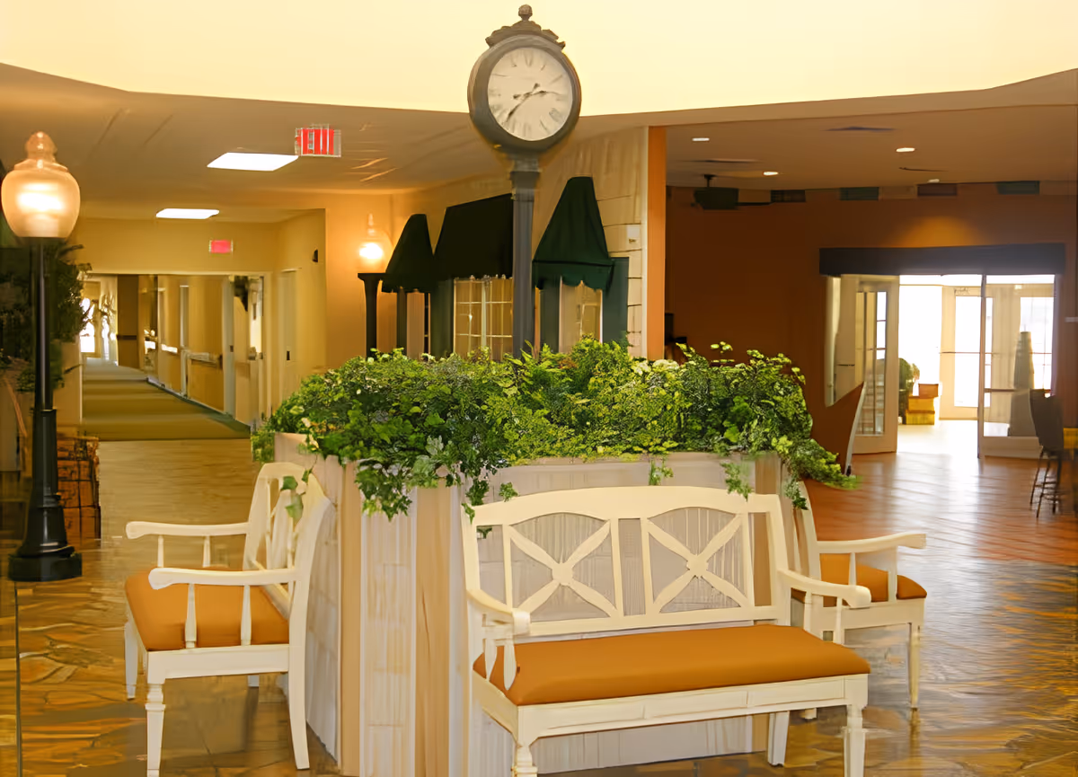 Indoor seating area with white wooden benches featuring orange cushions surrounding a planter filled with green plants. A vintage-style clock on a post stands behind the planter. The area is well-lit with ceiling lights and wall sconces, and a hallway extends to the left with multiple doors and exit signs visible.