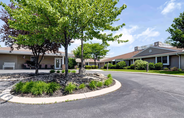 Exterior view of Celebration Villa of Altoona showing a circular driveway with landscaped greenery and trees in the center. The single-story building has a beige exterior with a brown roof and several windows. There are benches and chairs on the covered porch area near the entrance. The sky is partly cloudy with blue patches.