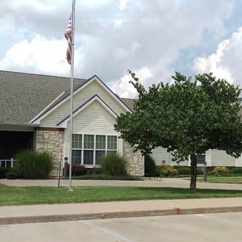 Exterior view of a single-story building with beige siding and stone accents, a flagpole with an American flag, a tree, and a well-maintained lawn under a partly cloudy sky.