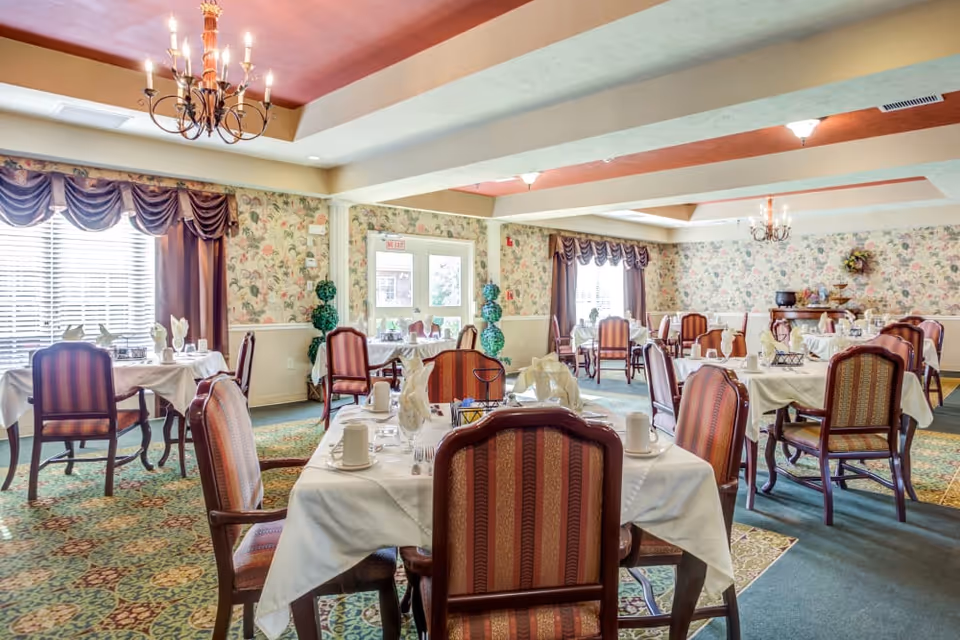 A dining room with multiple tables covered in white tablecloths, set with cups, napkins, and silverware. The room features patterned carpet, floral wallpaper, large windows with draped curtains, and chandeliers hanging from the ceiling.