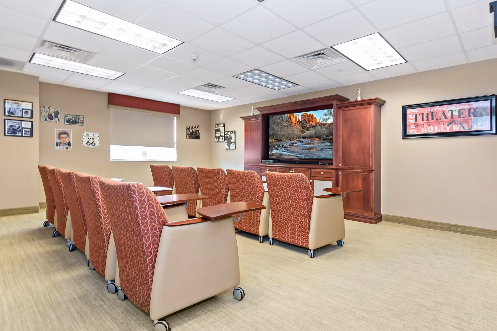 A small theater room with two rows of comfortable chairs with attached wooden trays facing a large flat-screen TV set in a wooden entertainment center. The walls are decorated with movie-themed posters and pictures, and the room has a beige carpet and neutral-colored walls.