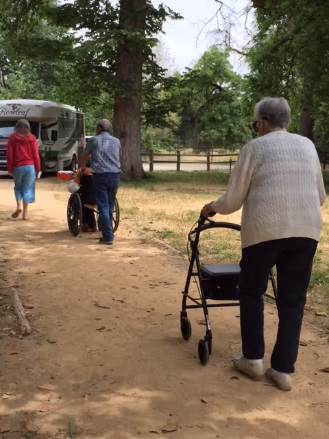 Three elderly individuals outdoors on a dirt path surrounded by trees. One person is using a walker, another is pushing a wheelchair, and a third person is walking ahead. A vehicle with the name 'Roseleaf' is parked nearby.
