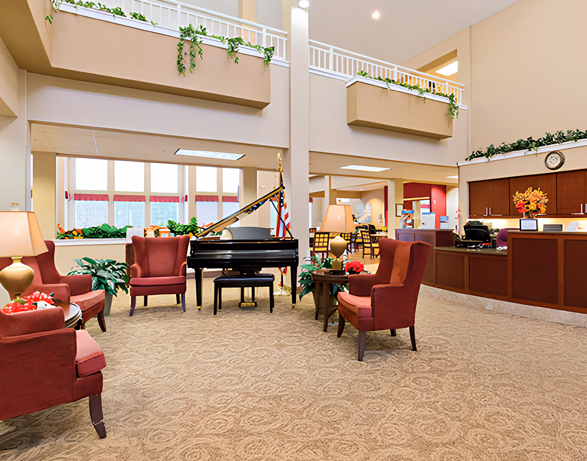 A spacious senior living facility common area with beige walls and carpet. The room features several red upholstered armchairs arranged around small tables with lamps and plants. A black grand piano with a bench is positioned near large windows that let in natural light. There is a reception desk with a clock above it and decorative plants on top. The area has a high ceiling with a balcony railing decorated with greenery.