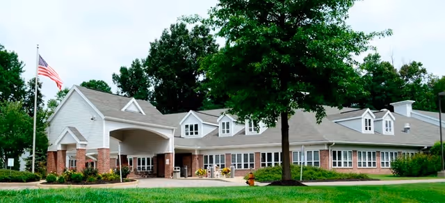 Exterior view of a single-story senior living facility building with a covered entrance, brick and white siding, multiple windows, a large tree in front, and an American flag on a flagpole to the left.