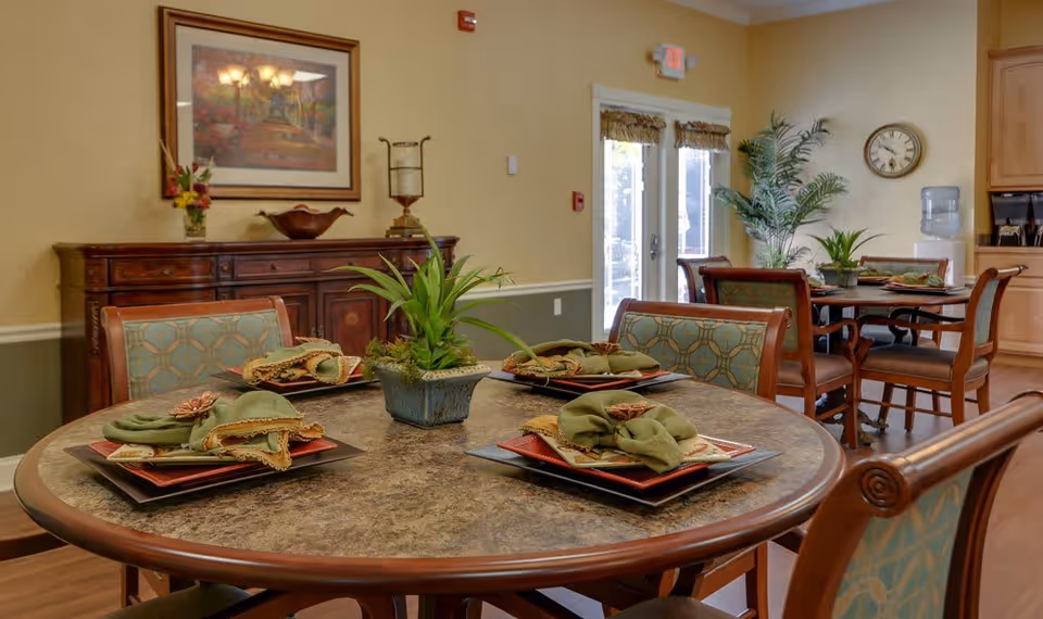 A dining area in a senior living facility with round tables set with plates, napkins, and decorative napkin rings. The room features wooden chairs with patterned upholstery, a sideboard with decorative items, a wall clock, potted plants, and double glass doors letting in natural light.