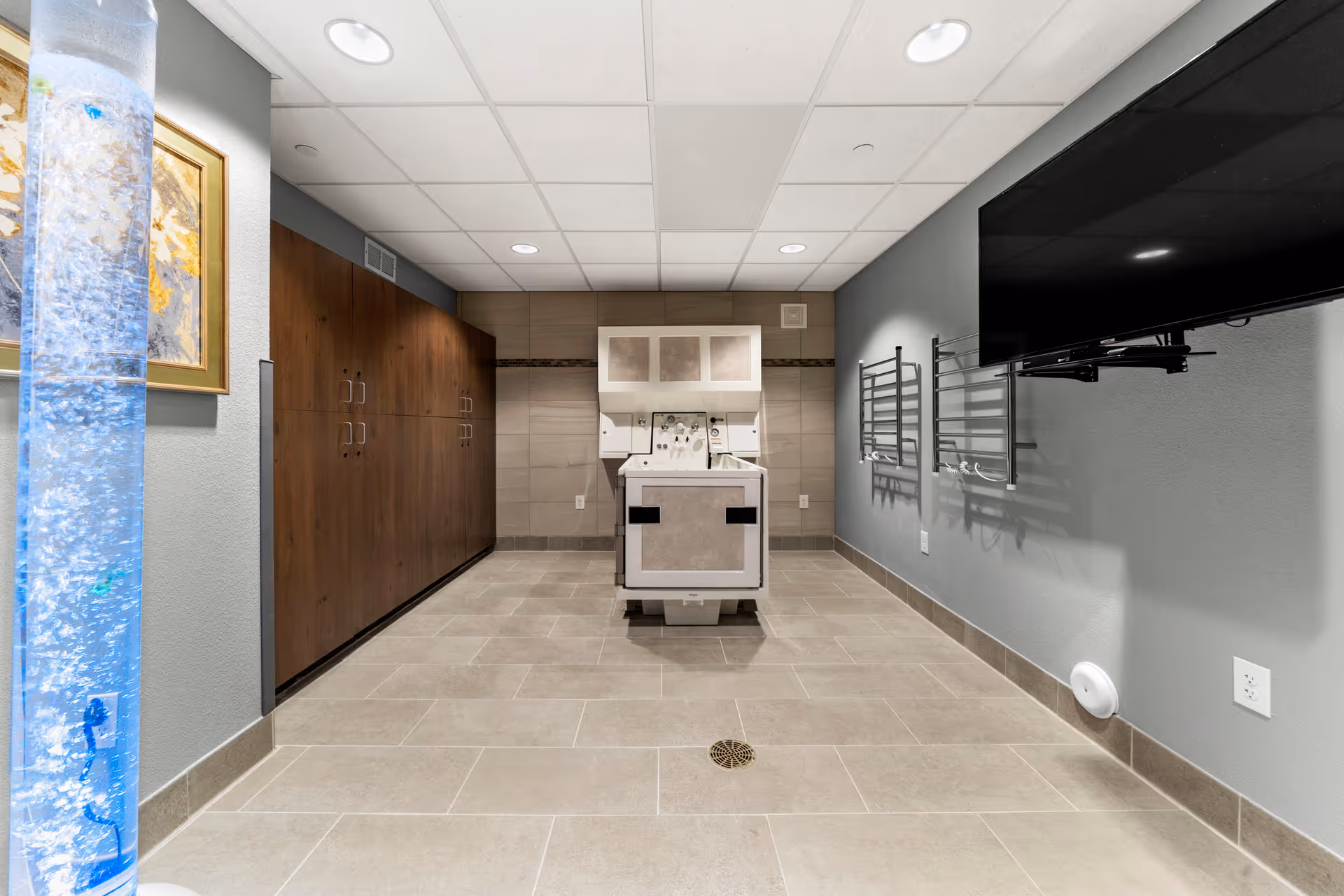 Tile-floored therapy room with a central patient lift/transfer chair, wall cabinets, a wall-mounted TV and a decorative bubble column.