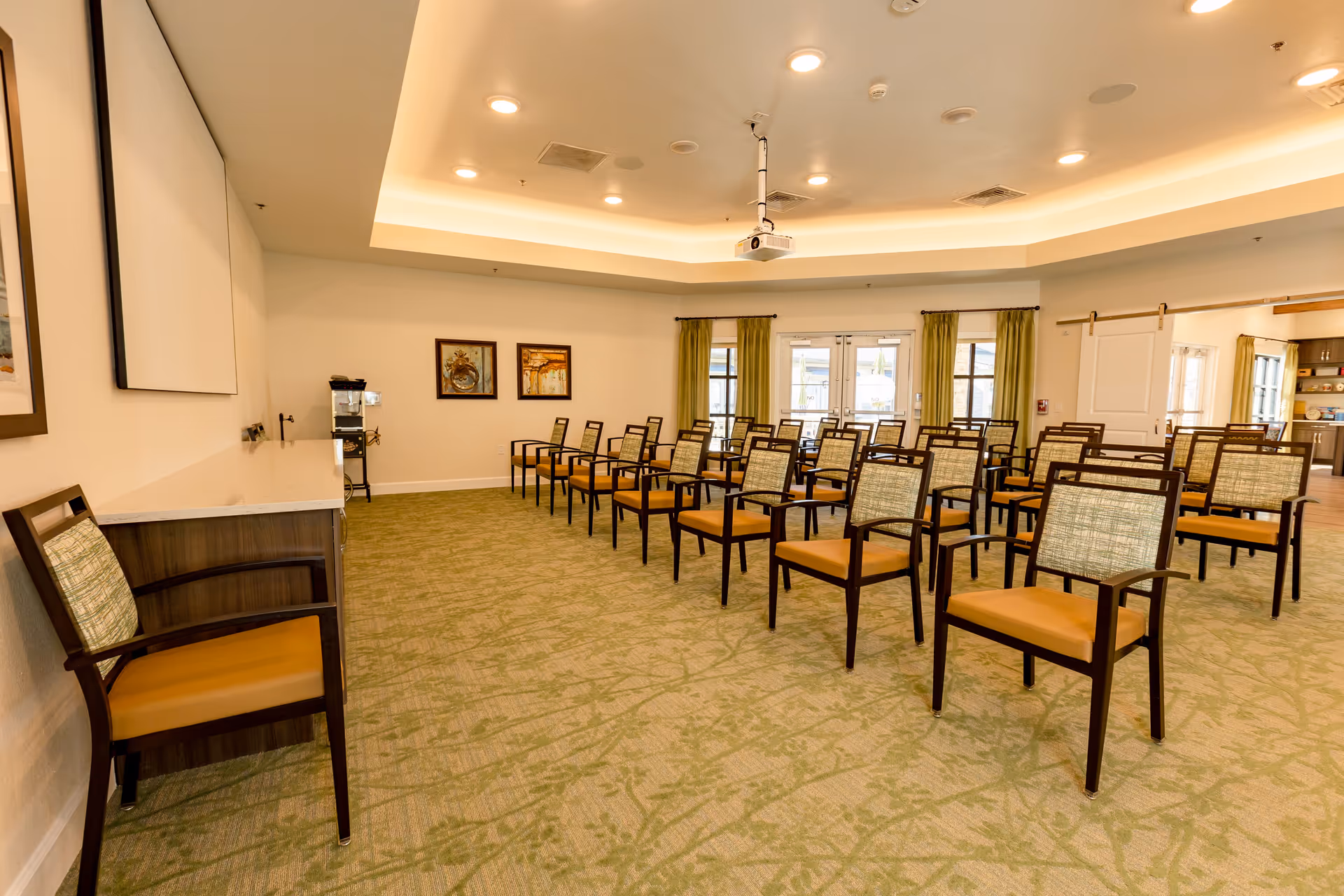 A well-lit meeting or presentation room with rows of wooden chairs featuring green patterned backs and tan seats arranged facing a blank projection screen on the wall. The room has green carpet with a subtle pattern, recessed ceiling lights, and large windows with green curtains allowing natural light. There are two framed artworks on the wall and a water dispenser in the corner.