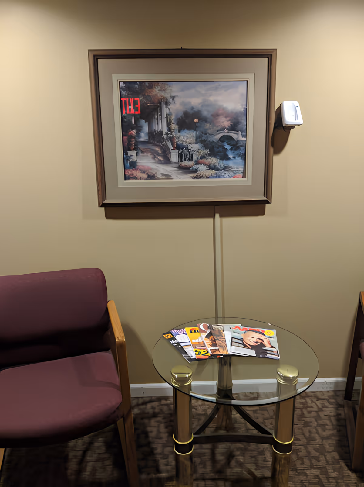 A small waiting area with two purple cushioned chairs and a round glass table between them. On the table, there are several magazines. A framed painting depicting a scenic outdoor scene hangs on the beige wall above the table. A white wall-mounted hand sanitizer dispenser is visible to the right of the painting.