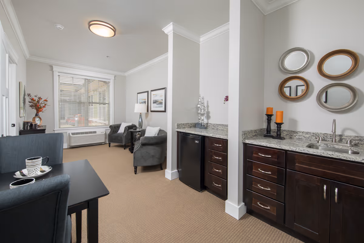 A cozy senior living room area with two gray armchairs, a small side table with a lamp, and two framed pictures on the wall. To the right, there is a kitchenette with dark wood cabinets, a granite countertop, a small sink, a mini refrigerator, and decorative mirrors and candles on the wall. A dining table with two chairs and a cup and saucer set is partially visible in the foreground. The room has beige carpet and a large window with blinds.