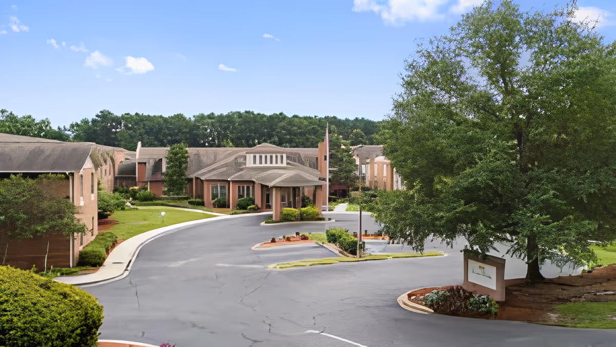 Exterior view of TerraBella Windsor Lake facility showing a curved driveway leading to the main entrance surrounded by landscaped greenery and trees under a clear blue sky.
