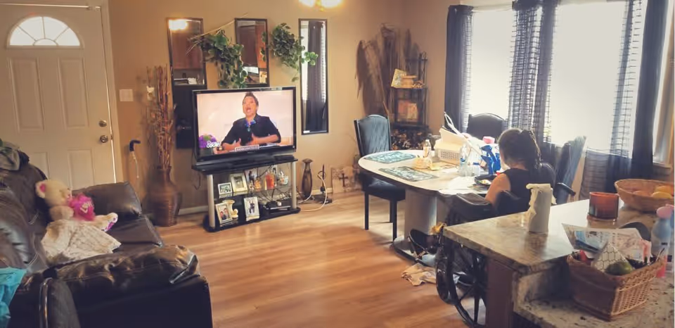 A cozy living room with wooden flooring, a brown leather couch with stuffed animals on it, a TV on a stand displaying a person speaking, and a woman sitting at a dining table near large windows with dark curtains. The room has various decorative items including plants, framed photos, and a basket on a countertop.