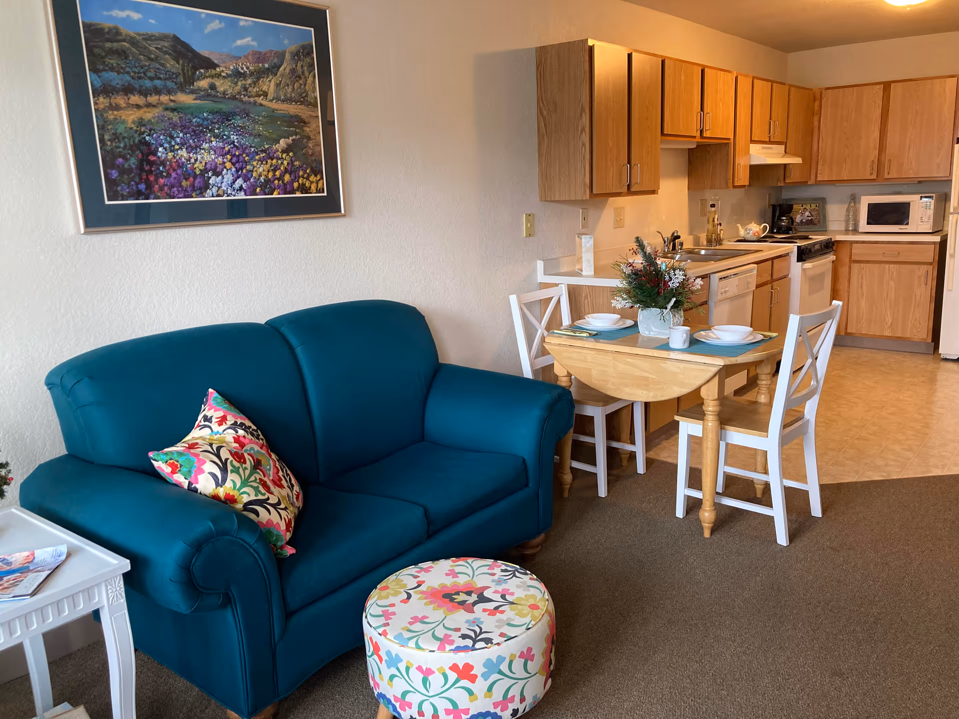 A cozy senior living space featuring a teal loveseat with a colorful floral pillow, a round floral-patterned ottoman, a small wooden dining table set for two with white chairs, and a kitchen area with wooden cabinets, a stove, microwave, and dishwasher. A landscape painting hangs on the wall above the loveseat.