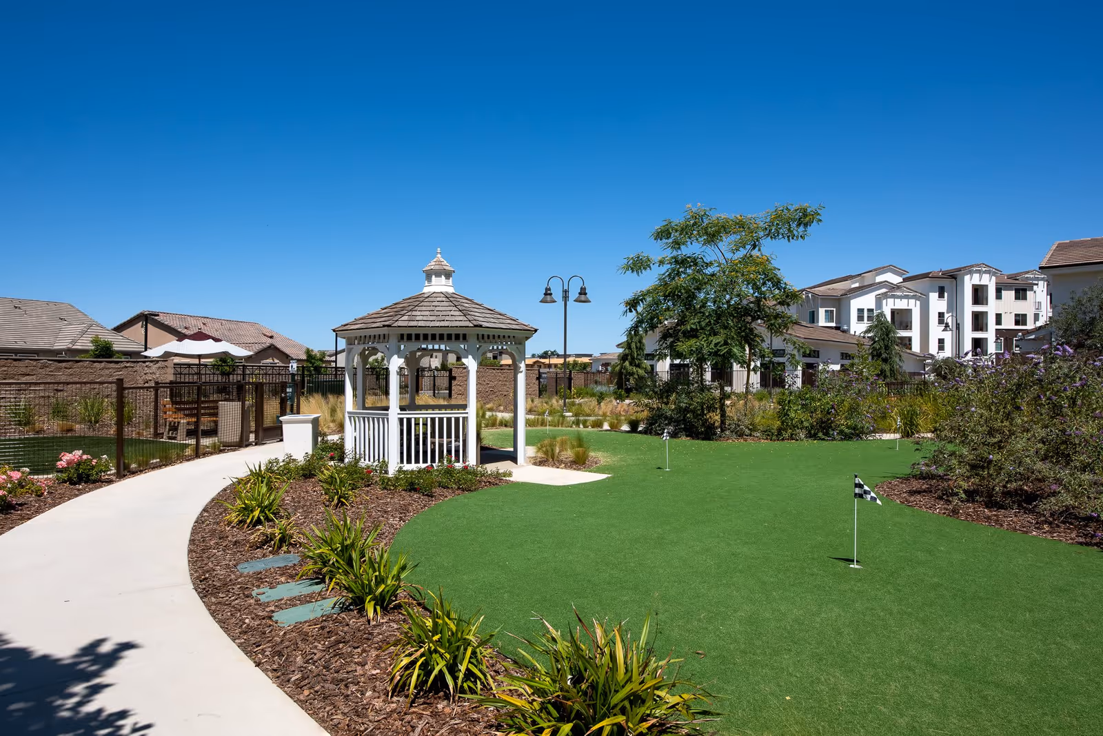 Outdoor garden area with a white gazebo, a putting green with small flags, landscaped plants, and a paved walkway under a clear blue sky. Residential buildings are visible in the background.