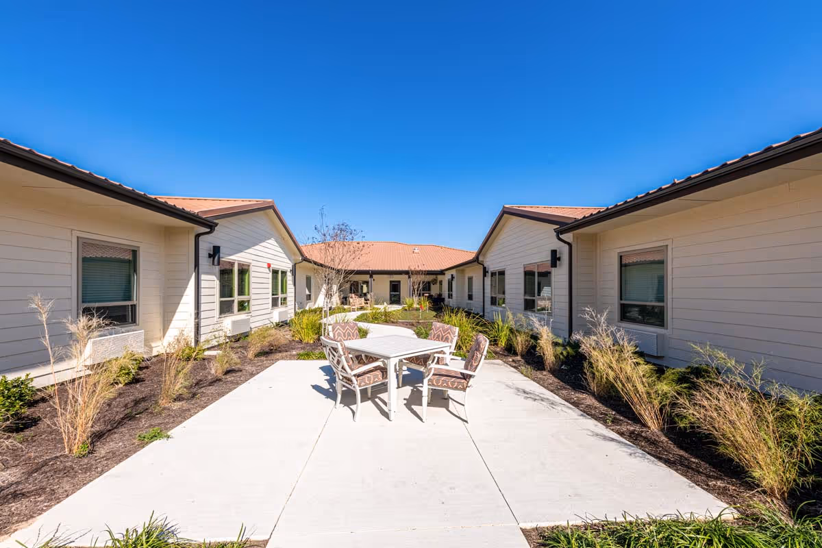 Outdoor courtyard area at The Philomena with a concrete patio, a table and four cushioned chairs in the center, surrounded by low landscaping and single-story buildings with beige siding and brown roofs under a clear blue sky.