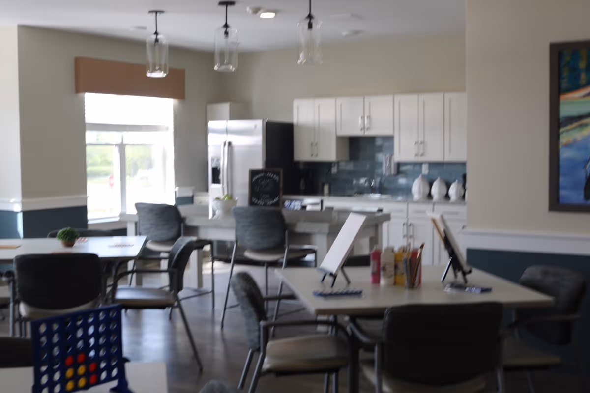 Interior view of a communal activity and dining area in a senior living facility. The room features several tables and chairs arranged for group activities, with art supplies and canvases on one table. In the background, there is a kitchen area with white cabinets, a stainless steel refrigerator, and a blue tiled backsplash. Large windows allow natural light to brighten the space.
