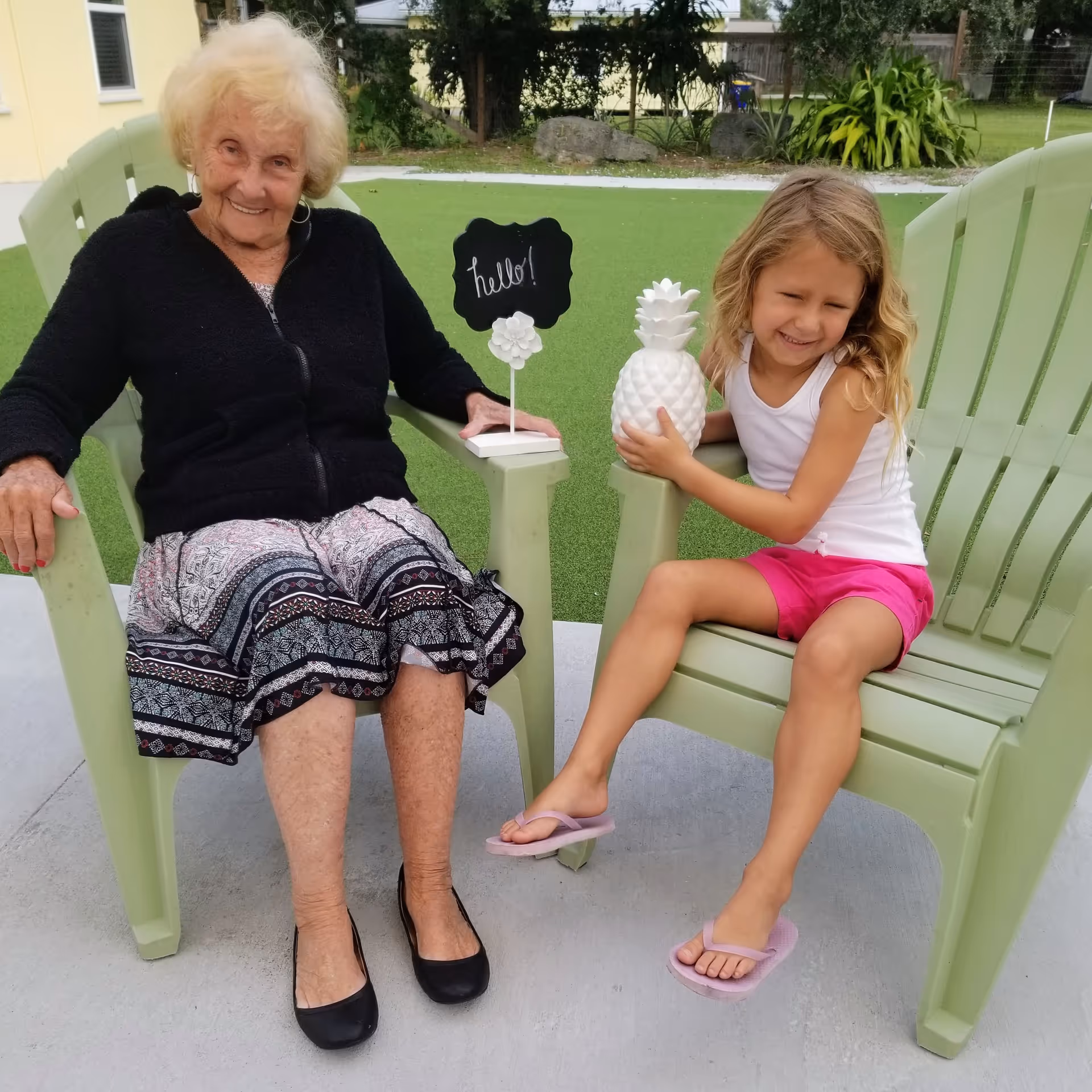 An elderly woman and a young girl sitting outdoors on light green Adirondack chairs. The elderly woman is wearing a black cardigan and patterned skirt, smiling at the camera. The young girl is wearing a white tank top and pink shorts, holding a white decorative pineapple and smiling with her eyes closed. Between them is a small blackboard sign with the word 'hello!' written on it. The background shows green artificial turf and some plants.