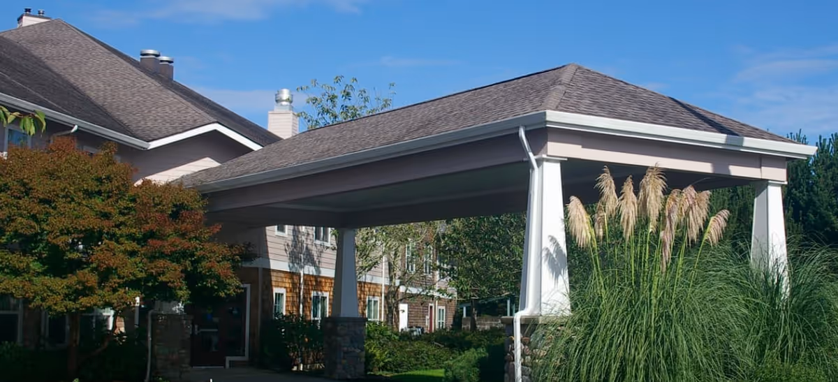 Exterior view of a senior living facility with a covered entrance supported by white pillars and stone bases, surrounded by green shrubs and tall ornamental grasses under a clear blue sky.