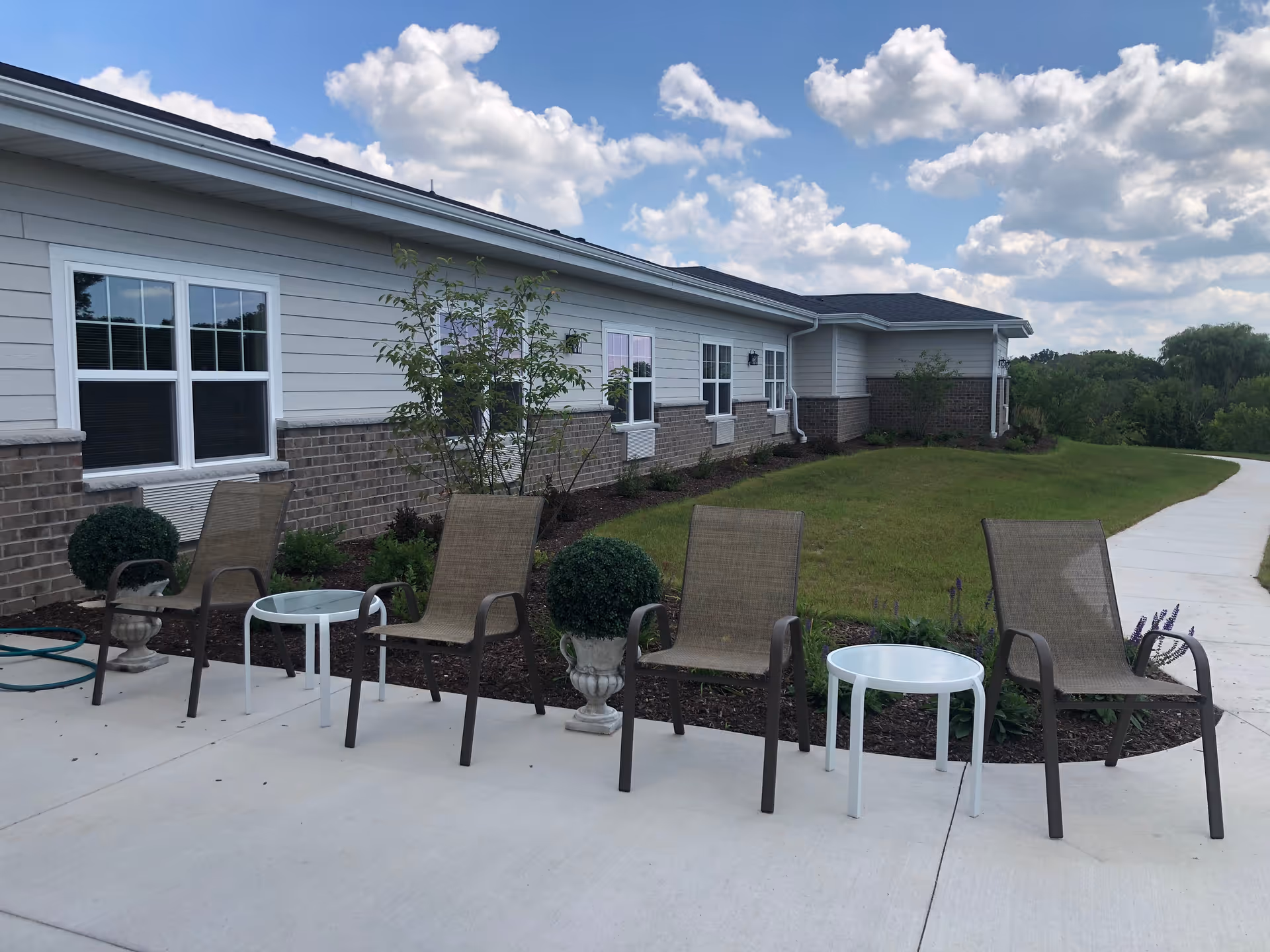 Outdoor seating area with several chairs and small tables along a walkway in front of a single-story senior living building under a partly cloudy sky.