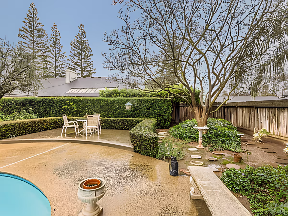 A wet backyard patio with a small pool, a table and chairs, trimmed hedges, a tree, and a stepping-stone garden path.