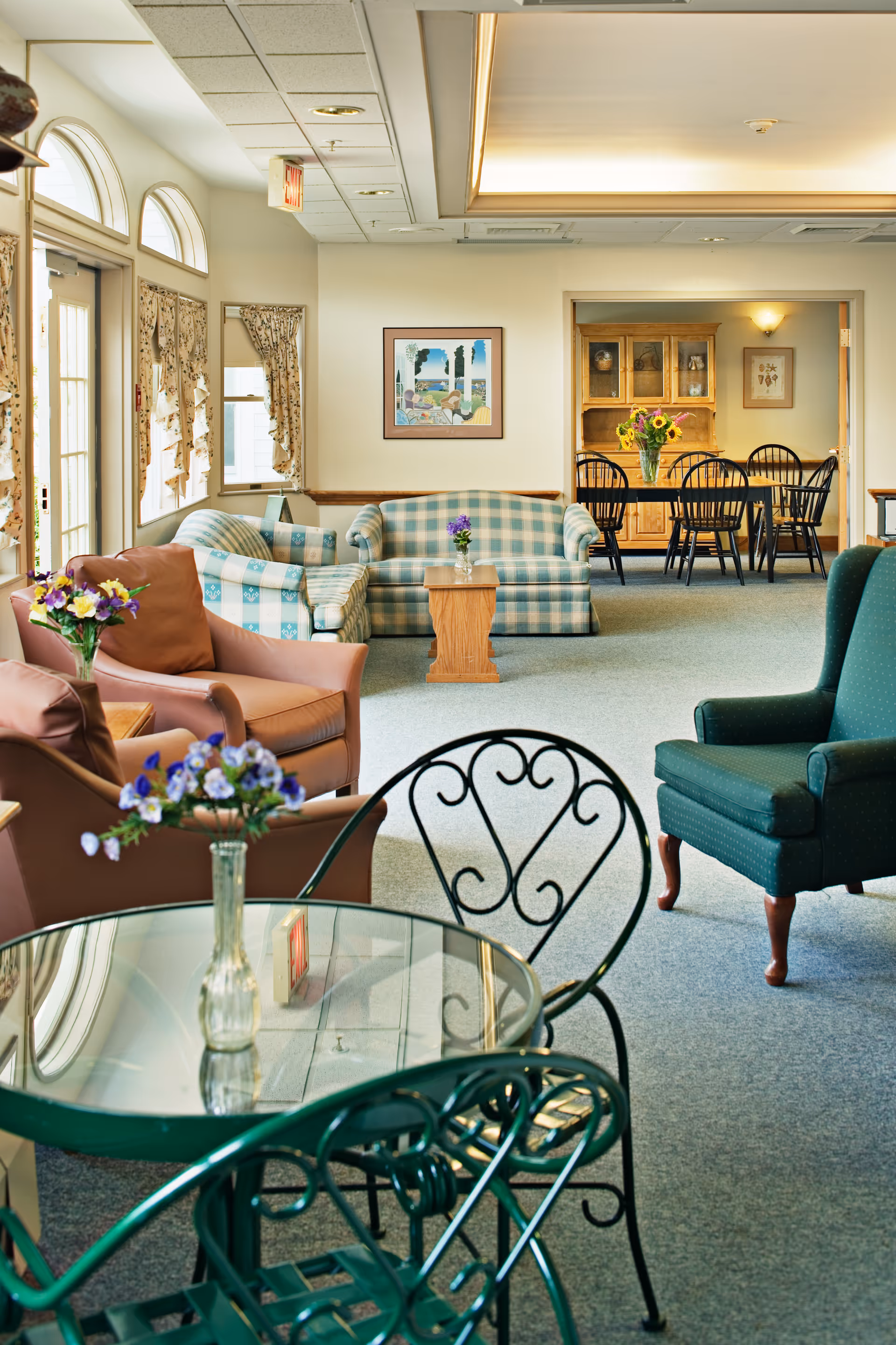A cozy senior living facility common area with various seating options including a green armchair, a brown leather armchair, and a blue and white checkered sofa. There is a glass-top table with a vase of purple flowers in the foreground and a wooden coffee table with a small flower vase in the middle. In the background, there is a dining area with a wooden cabinet and a dining table with black chairs, decorated with a vase of sunflowers. The room has large windows with floral curtains and soft lighting.