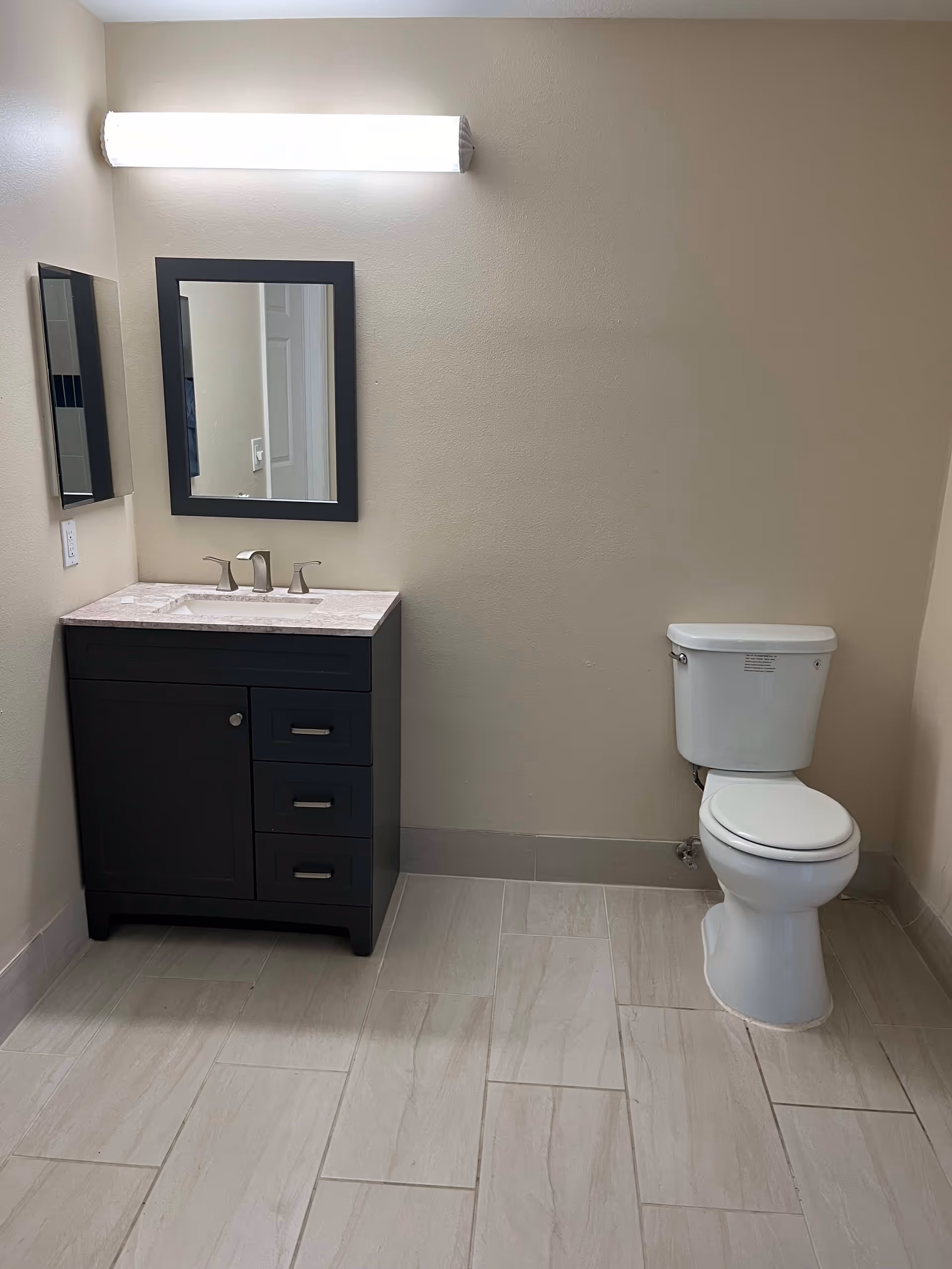 A clean bathroom with a dark wood vanity featuring a marble countertop and a rectangular mirror above it. To the right of the vanity is a white toilet. The floor is tiled with light-colored rectangular tiles, and the walls are painted beige. A bright horizontal light fixture is mounted above the mirror.