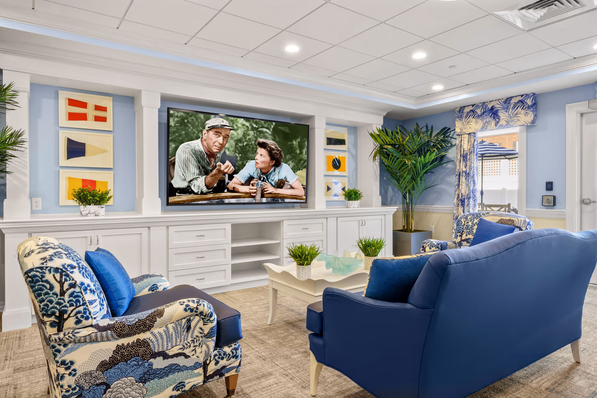 A cozy living room with blue and patterned armchairs and a blue sofa arranged around a white coffee table with small green plants. A large flat-screen TV is mounted on a white built-in cabinet wall, displaying a black and white movie scene. The room has light blue walls, decorative framed artwork, a large potted plant, and a window with blue patterned curtains letting in natural light.