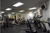 Interior view of a fitness room with various exercise equipment including treadmills, stationary bikes, and weight machines. The room has white walls, a black floor, fluorescent ceiling lights, and a wall-mounted television.