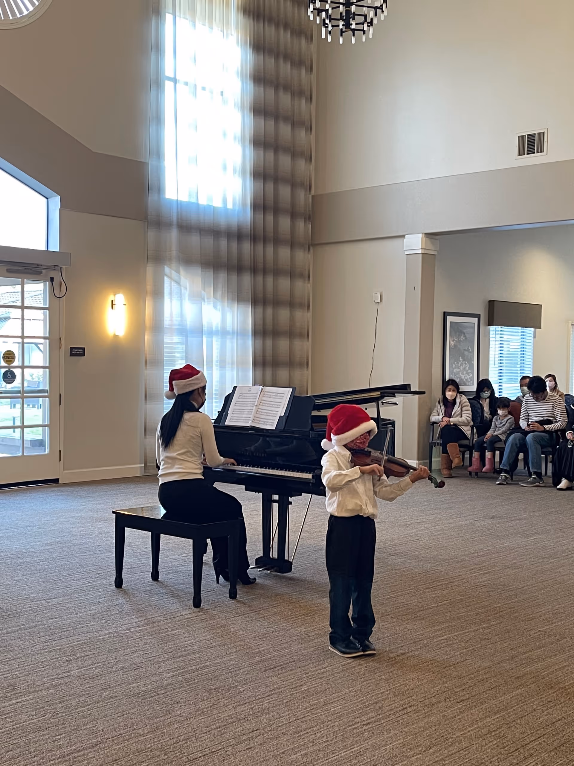 A woman wearing a Santa hat plays the piano while a young boy, also wearing a Santa hat, plays the violin in a spacious room with large windows and high ceilings. Several people are seated in the background watching the performance.