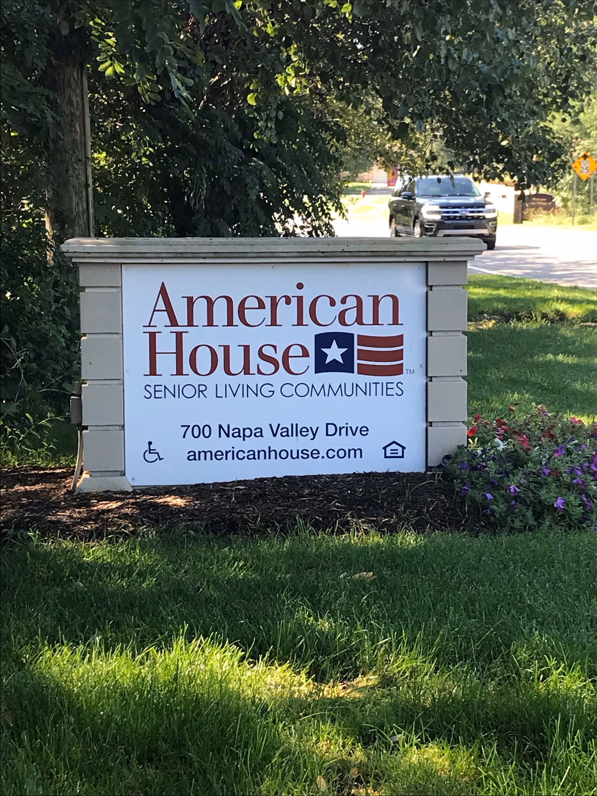 Outdoor sign for American House Senior Living Communities located at 700 Napa Valley Drive with the website americanhouse.com, surrounded by grass, flowers, and trees, with a road and a vehicle in the background.