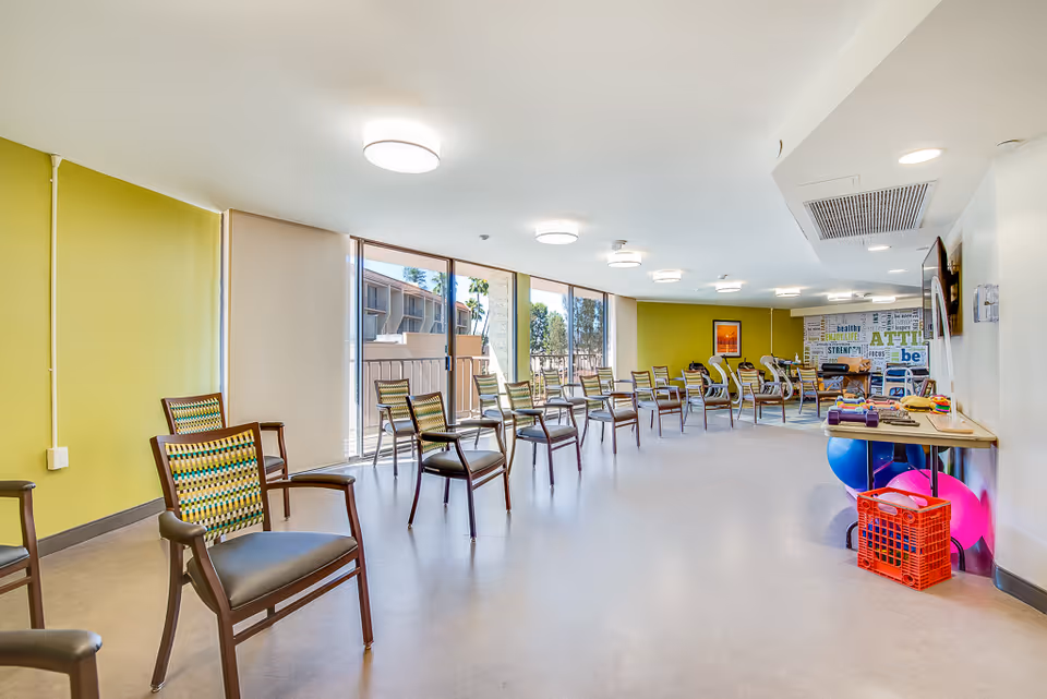 A spacious, well-lit room with multiple chairs arranged in rows facing a wall with motivational words. Large windows let in natural light, and exercise equipment including colorful exercise balls and weights are visible on the right side of the room.