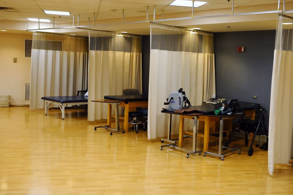 A rehabilitation or physical therapy room with wooden floors, several treatment tables separated by beige privacy curtains, chairs, and exercise equipment including a pedal exerciser and small weights.