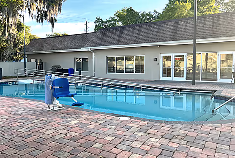 Outdoor swimming pool area with a pool lift chair for accessibility, surrounded by a brick-paved deck. In the background, there is a single-story building with large windows and glass doors. Trees and a clear sky are visible behind the building.