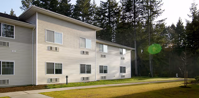 Exterior side of a two-story light-colored senior living building with multiple windows, a lawn, walkway and tall trees in the background.