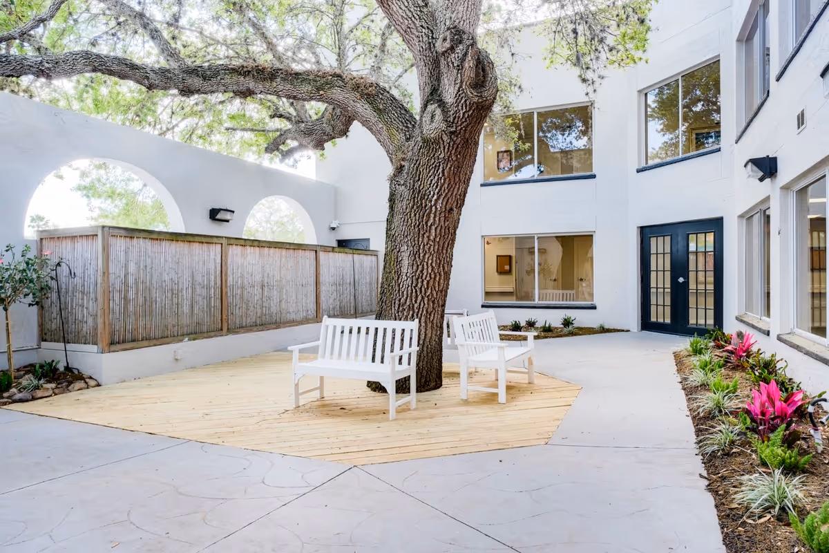 Outdoor courtyard area with a large tree in the center surrounded by a wooden deck. Two white benches are placed on the deck around the tree. The courtyard is enclosed by white walls with arched openings and windows, and there are plants and flowers along the edges.