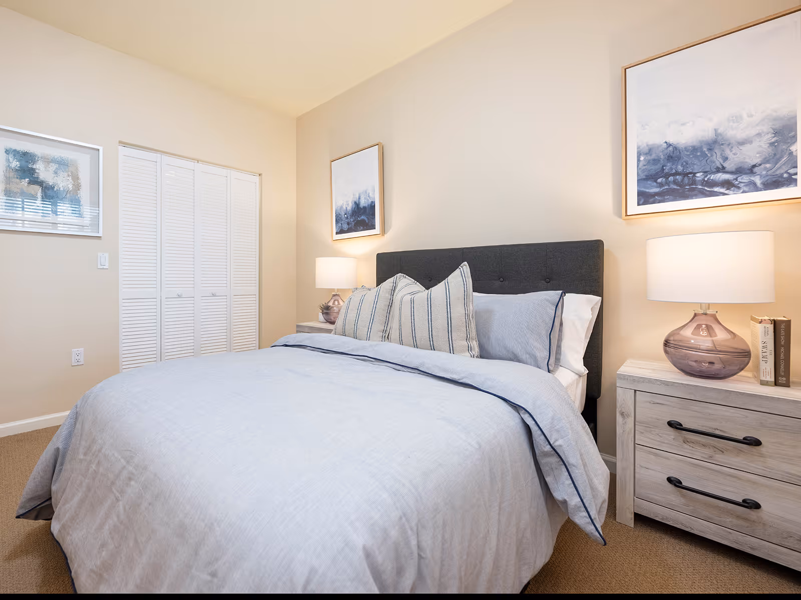 A neatly made bed with light blue bedding and three striped pillows in a bedroom. There are two nightstands on either side of the bed, each with a lamp and some books. The walls are beige with two abstract paintings hanging above the bed and near the closet. The closet has white louvered doors.