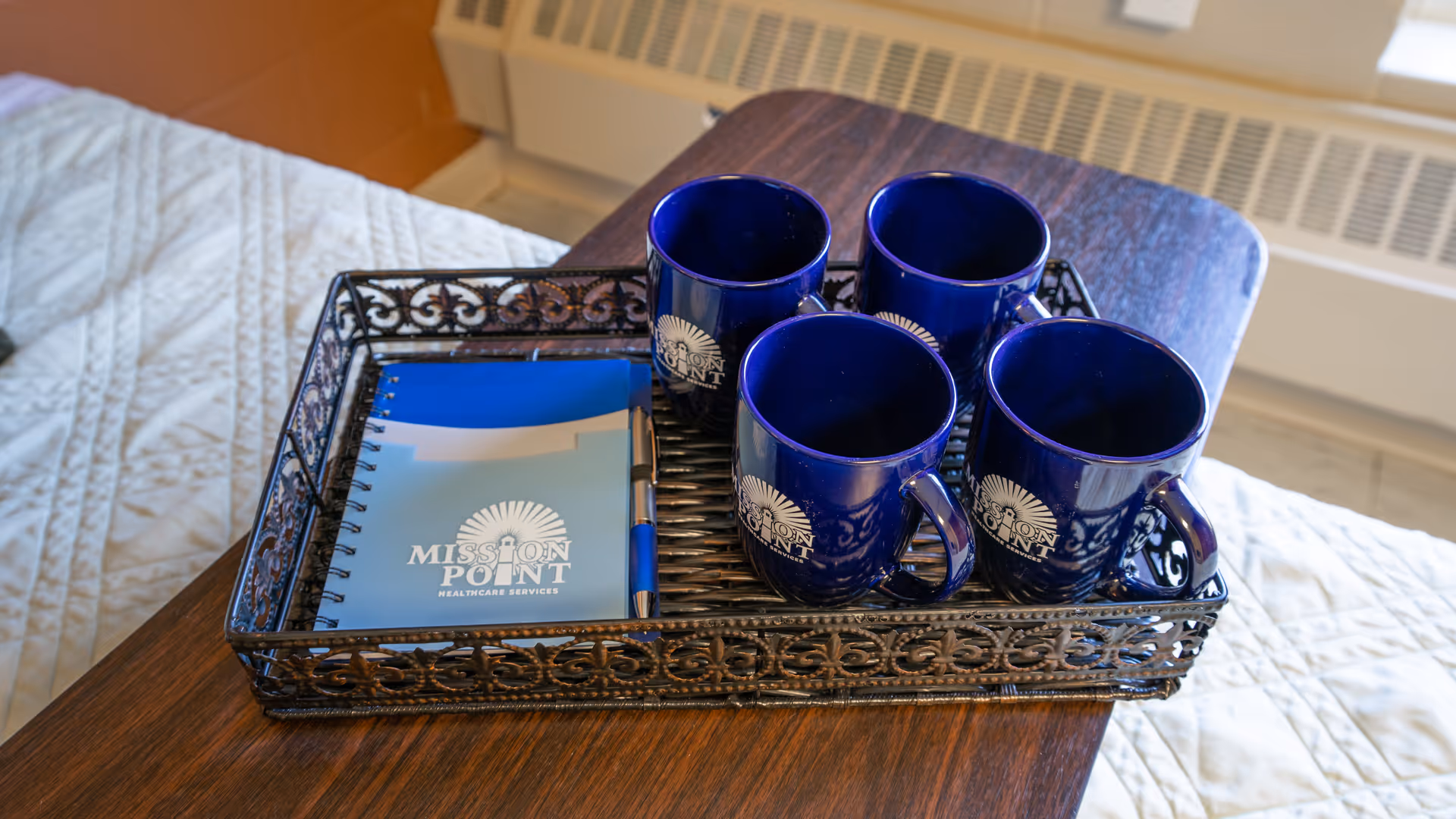 Metal tray on a wooden bedside table holding four blue "Mission Point" mugs and a matching notebook with a quilted bed visible in the background.
