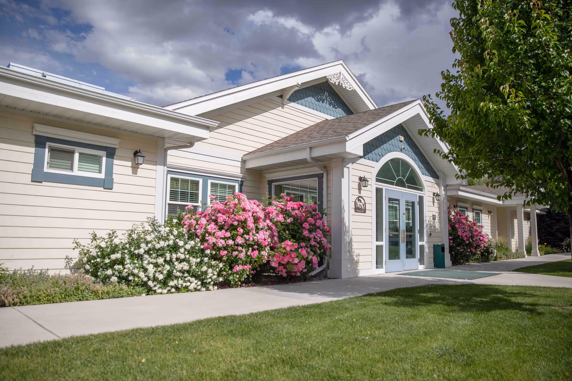 Exterior view of a senior living facility building with beige siding and blue trim. The entrance features double glass doors with an arched window above. There are blooming pink and white flowers along the walkway and a green lawn in the foreground. A tree is visible on the right side under a partly cloudy sky.