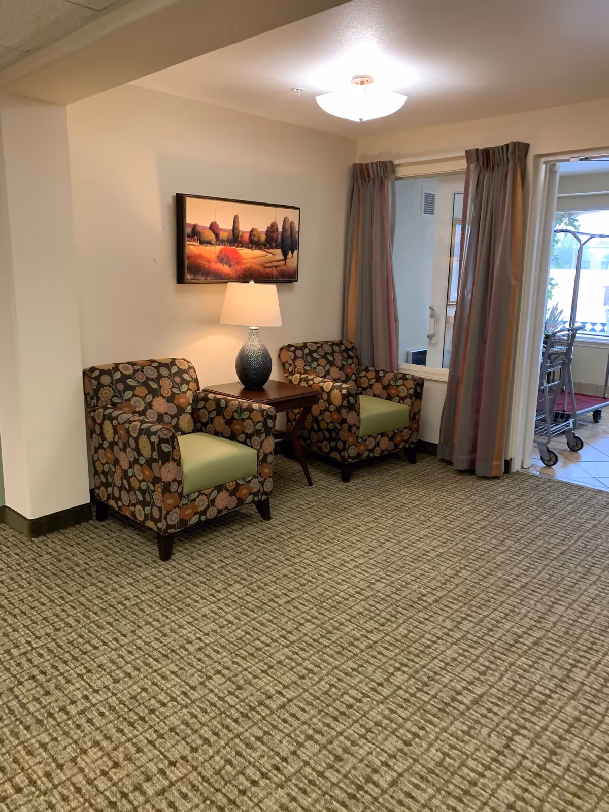 A cozy seating area with two floral-patterned armchairs with green cushions, separated by a wooden side table holding a decorative lamp. Above the chairs hangs a landscape painting. To the right, there is a window with curtains and a door leading to another room with tiled flooring and a luggage cart.