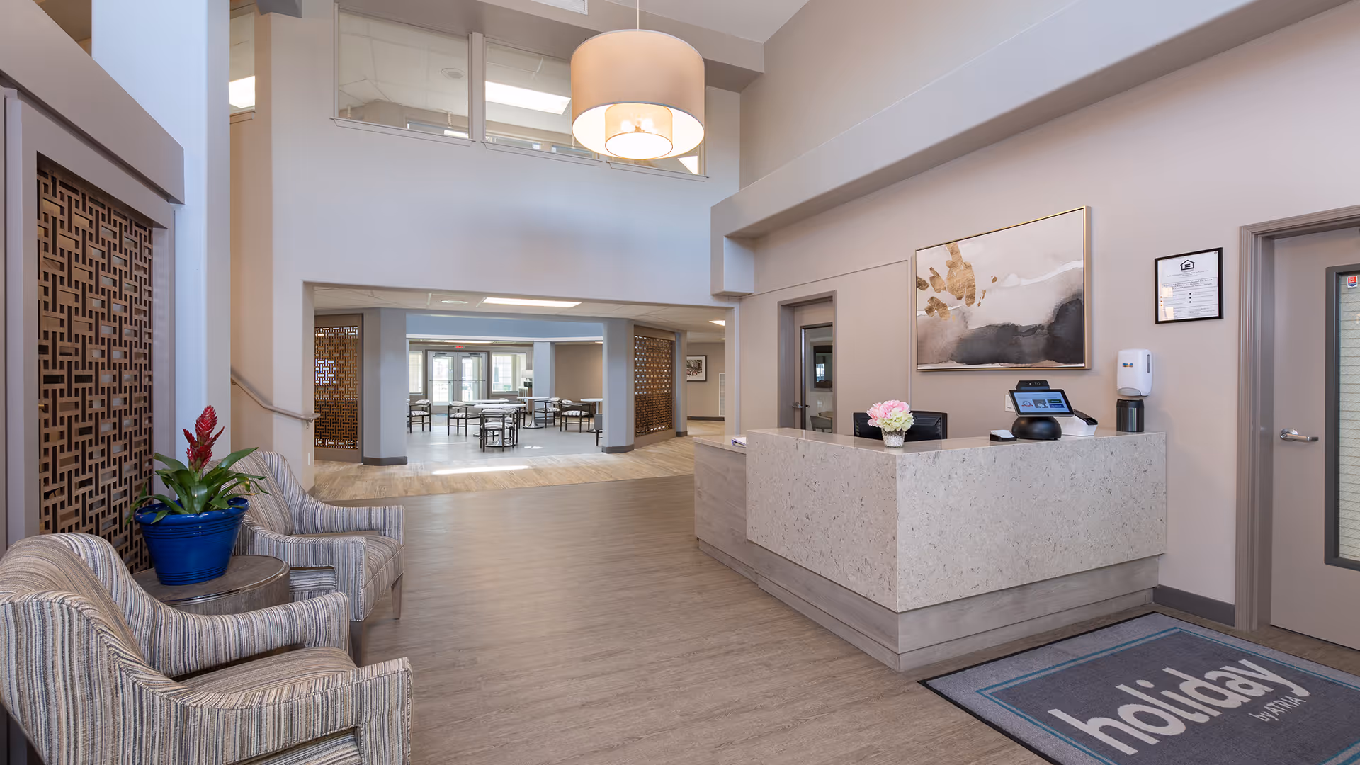 Reception area of Highland Trail | Sky Active Living featuring a modern front desk with a computer and a vase of flowers, two striped armchairs with a small round table and a potted plant, a large hanging light fixture, and a view into a dining area with tables and chairs.