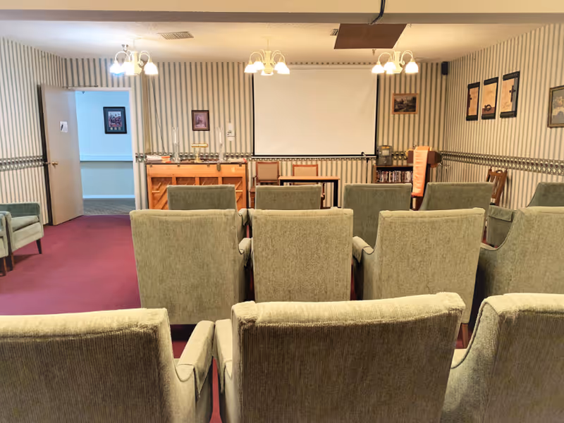 A small meeting or presentation room with several rows of green upholstered chairs facing a white projection screen. The room has striped wallpaper, framed pictures on the walls, a piano, and a small table with chairs at the front. Overhead lights hang from the ceiling, and an open door leads to another room.