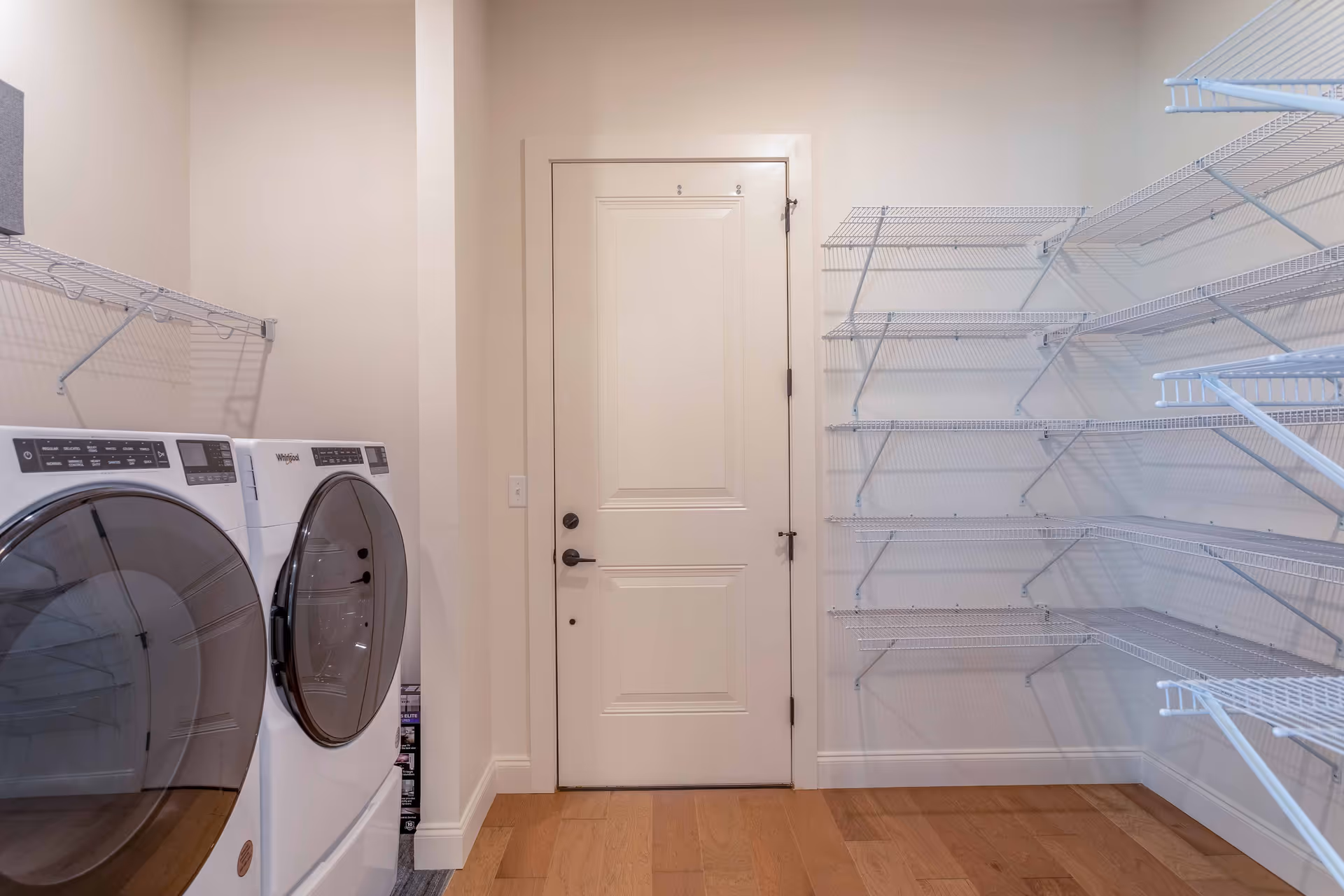 Laundry room with a white front-loading washer and dryer on the left side, white wire shelving on the right wall, and a closed white door in the center. The floor is wooden and the walls are painted light beige.