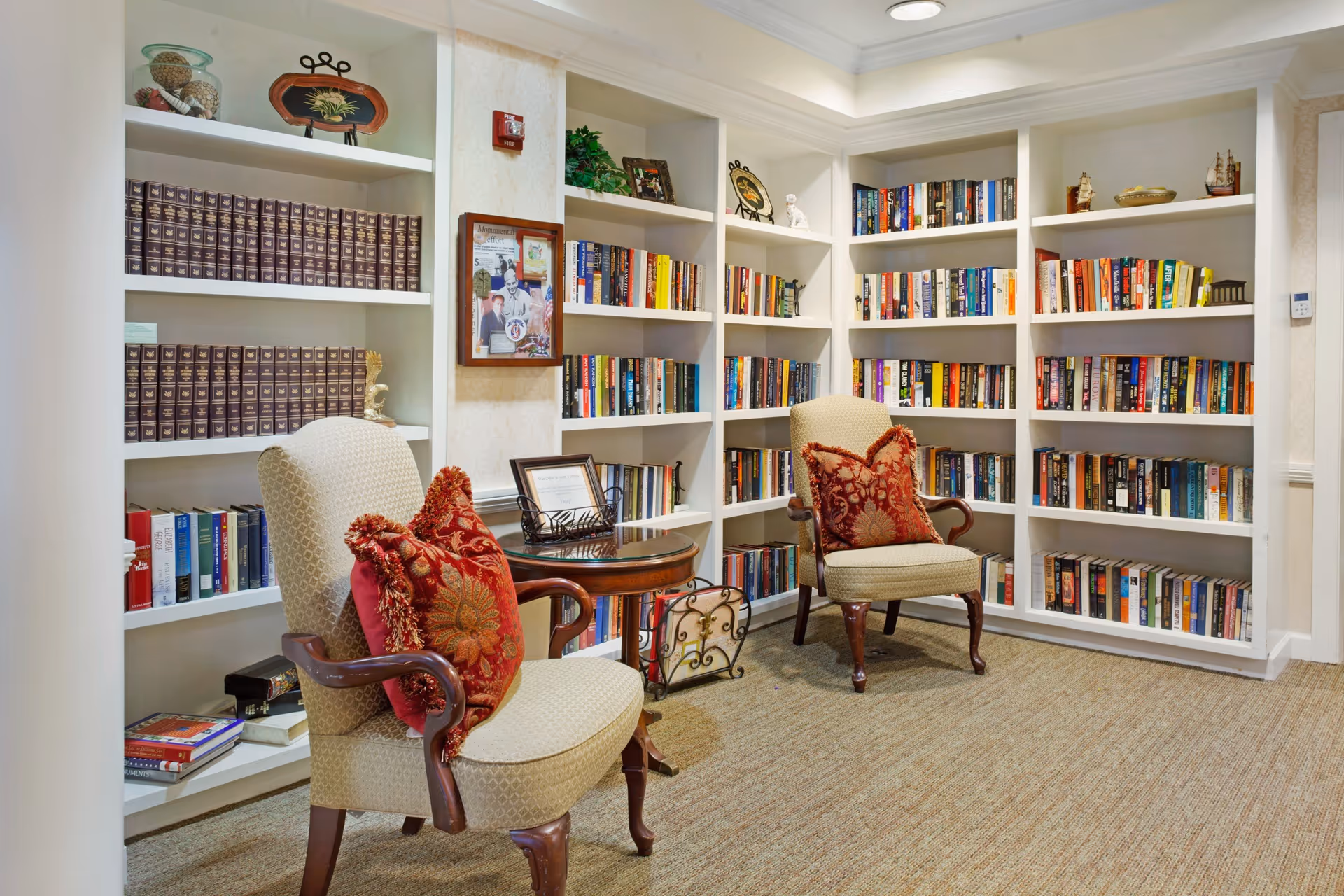 A cozy reading nook in a senior living facility featuring two upholstered armchairs with red decorative pillows, a round wooden side table between them, and built-in white bookshelves filled with books and decorative items along the walls.