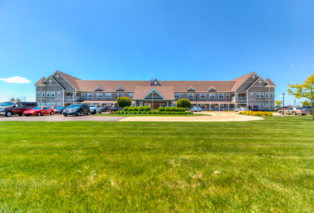 Front exterior of a large two-story senior living facility with a manicured lawn, driveway, and parked cars under a clear blue sky.