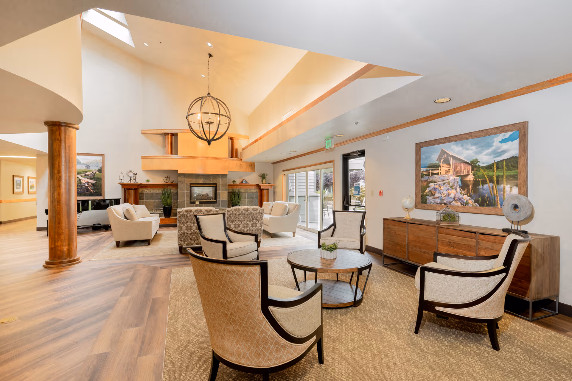 A spacious and well-lit living room area in a senior living facility featuring a high vaulted ceiling with a large modern chandelier. The room has a fireplace with a wooden mantel, several upholstered armchairs arranged around a round coffee table, and a wooden sideboard with decorative items and a large framed landscape painting above it. Large windows and glass doors allow natural light to fill the space.