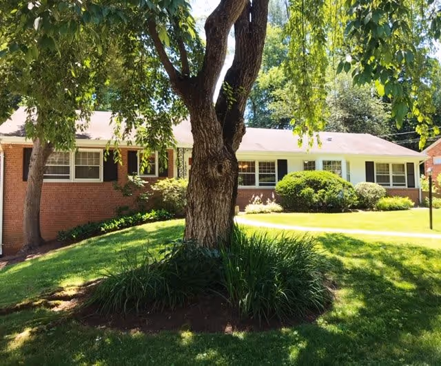 Single-story brick building with white window frames and black shutters, surrounded by green grass, bushes, and a large tree in the foreground under a sunny sky.